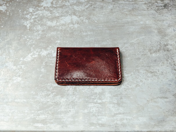 A dark brown leather wallet with white stitching along its edges is placed on a textured, gray surface. The wallet's surface appears glossy with some natural creases visible.