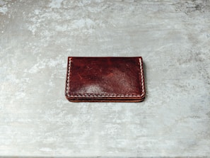 A dark brown leather wallet with white stitching along its edges is placed on a textured, gray surface. The wallet's surface appears glossy with some natural creases visible.