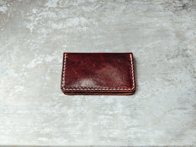 A dark brown leather wallet with white stitching along its edges is placed on a textured, gray surface. The wallet's surface appears glossy with some natural creases visible.