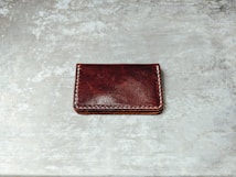 A dark brown leather wallet with white stitching along its edges is placed on a textured, gray surface. The wallet's surface appears glossy with some natural creases visible.
