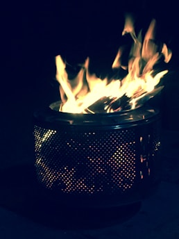 A fire burning inside a metal drum with holes, creating a glowing effect. The flames are vivid, casting a warm light against a dark backdrop.