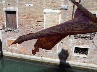 A decorative purple and gold fabric banner with intricate floral patterns is hanging over a narrow canal. The background features a weathered brick wall with two windows, one having a closed wooden shutter and the other covered with decorative iron bars. The reflection of the banner is visible in the water below.