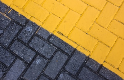 A close-up view of a paved surface with interlocking bricks. The bricks are arranged in two distinct sections; one side is painted yellow and the other is gray, creating a diagonal division.