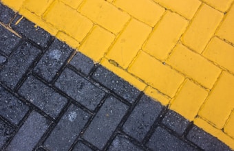 A close-up view of a paved surface with interlocking bricks. The bricks are arranged in two distinct sections; one side is painted yellow and the other is gray, creating a diagonal division.
