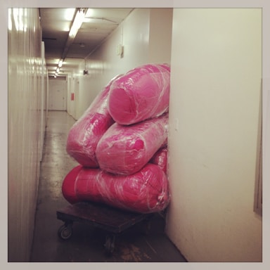 A narrow hallway with industrial lighting contains a wheeled cart loaded with large, wrapped pink objects that are stacked against the wall. The corridor appears to have cream-colored walls and a shiny floor.