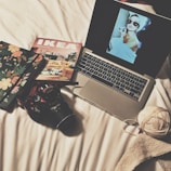 A cozy, creative setup on a white bed includes a laptop displaying a vintage-style image of a woman with large sunglasses and a towel on her head applying lipstick. Nearby, a floral-patterned notebook, an IKEA catalog, a DSLR camera, a ball of yarn, and a piece of knitted fabric are arranged neatly.