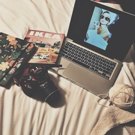 A cozy, creative setup on a white bed includes a laptop displaying a vintage-style image of a woman with large sunglasses and a towel on her head applying lipstick. Nearby, a floral-patterned notebook, an IKEA catalog, a DSLR camera, a ball of yarn, and a piece of knitted fabric are arranged neatly.