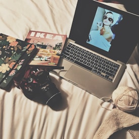A cozy, creative setup on a white bed includes a laptop displaying a vintage-style image of a woman with large sunglasses and a towel on her head applying lipstick. Nearby, a floral-patterned notebook, an IKEA catalog, a DSLR camera, a ball of yarn, and a piece of knitted fabric are arranged neatly.