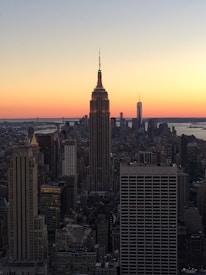 A city skyline at sunset with the Empire State Building prominently featured in the center. The horizon glows with warm shades of orange and pink as the sun sets, casting a gentle light over the numerous skyscrapers that populate the scene. The architecture is clearly defined against the colorful backdrop, with water visible in the distance.