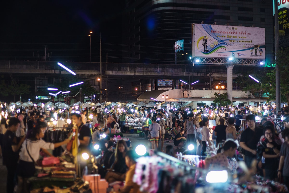 A vibrant Thai street scene with groups of people gathered at food stalls and tables, enjoying the bustling evening market atmosphere under warm lighting.. Photo by Adam Dore on Unsplash.