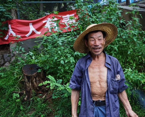 An elderly man stands outdoors wearing a straw hat and an open shirt, smiling. Behind him is lush green foliage and a red banner with white text in a non-Latin script.