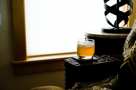 A cozy indoor setting featuring a glass filled with a cold, amber beverage resting on a wooden surface. Soft lighting filters through a window, casting a warm glow across the scene. A decorative lamp and a plush cushion add to the comfortable ambiance.