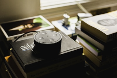 A small smart speaker is positioned on top of a stack of books, with the speaker's distinctive ring and buttons clearly visible. Surrounding the speaker, several other books and albums are scattered, creating a cluttered yet cozy atmosphere. The lighting in the room is dim, with muted colors that suggest a relaxed or contemplative setting.