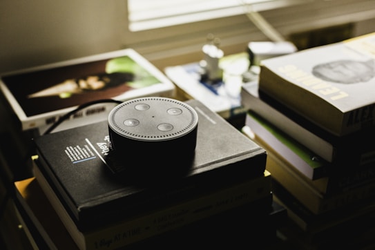 A small smart speaker is positioned on top of a stack of books, with the speaker's distinctive ring and buttons clearly visible. Surrounding the speaker, several other books and albums are scattered, creating a cluttered yet cozy atmosphere. The lighting in the room is dim, with muted colors that suggest a relaxed or contemplative setting.