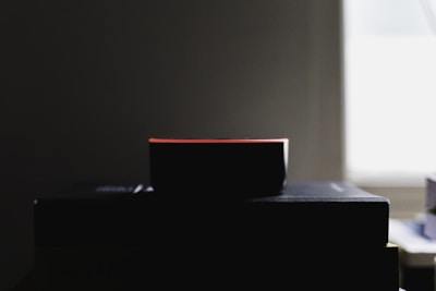 A dark object, likely a smart speaker or small electronic device, is silhouetted against a dimly lit background. The object sits on top of a stack of books or rectangular surfaces near a window emitting soft light.