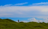 A solitary person stands on a gently rolling green hill under a vast blue sky filled with scattered clouds. The horizon is open and expansive, creating a sense of freedom and space.