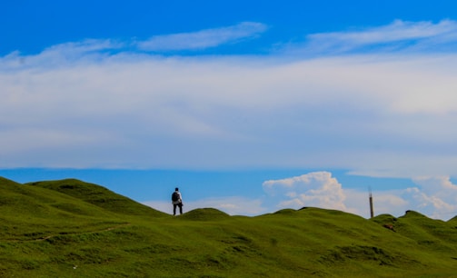 A solitary person stands on a gently rolling green hill under a vast blue sky filled with scattered clouds. The horizon is open and expansive, creating a sense of freedom and space.
