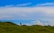 A solitary person stands on a gently rolling green hill under a vast blue sky filled with scattered clouds. The horizon is open and expansive, creating a sense of freedom and space.