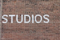 Large white letters spelling 'STUDIOS' are painted on a textured red brick wall, with the bricks arranged in a standard horizontal pattern.