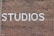 Large white letters spelling 'STUDIOS' are painted on a textured red brick wall, with the bricks arranged in a standard horizontal pattern.