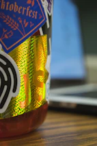 A close-up view of a colorful bottle covered with various stickers. The stickers include a blue one with 'Hacktoberfest' written in orange text, and a black sticker with abstract design elements. There's a reflective, holographic sticker with 'JS' prominently displayed. The background shows a blurred laptop, indicating a tech-related setting.