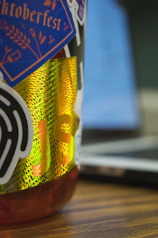 A close-up view of a colorful bottle covered with various stickers. The stickers include a blue one with 'Hacktoberfest' written in orange text, and a black sticker with abstract design elements. There's a reflective, holographic sticker with 'JS' prominently displayed. The background shows a blurred laptop, indicating a tech-related setting.