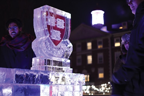 A nighttime scene featuring an intricately carved ice sculpture bearing a red and white emblem with the word 'Harvard'. The sculpture is illuminated, making it a focal point amidst the surrounding darkness. In the background, there are buildings and decorative lights, giving a festive atmosphere. Several people stand nearby, dressed warmly, indicating cold weather.