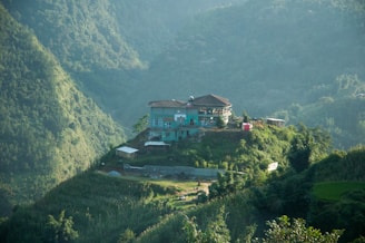 A house situated atop a small hill surrounded by lush green mountains, with dense vegetation covering the slopes. The house is blue and features multiple stories and a flat tiled roof. Surrounding the house, there are patches of greenery and small structures.