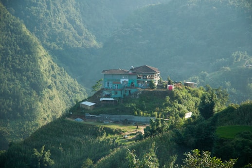 A house situated atop a small hill surrounded by lush green mountains, with dense vegetation covering the slopes. The house is blue and features multiple stories and a flat tiled roof. Surrounding the house, there are patches of greenery and small structures.