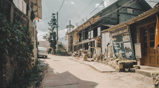A quiet street scene featuring rustic buildings with signage that offers massage and herb bath services. There are several potted plants lining the sidewalk, and a white car is parked along the left. The setting has an old-fashioned and somewhat rural feel, with visible utility lines overhead and a partially cloudy sky.