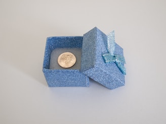 A small, glittery blue gift box with a lid adorned by a matching ribbon is open, revealing a single coin inside.