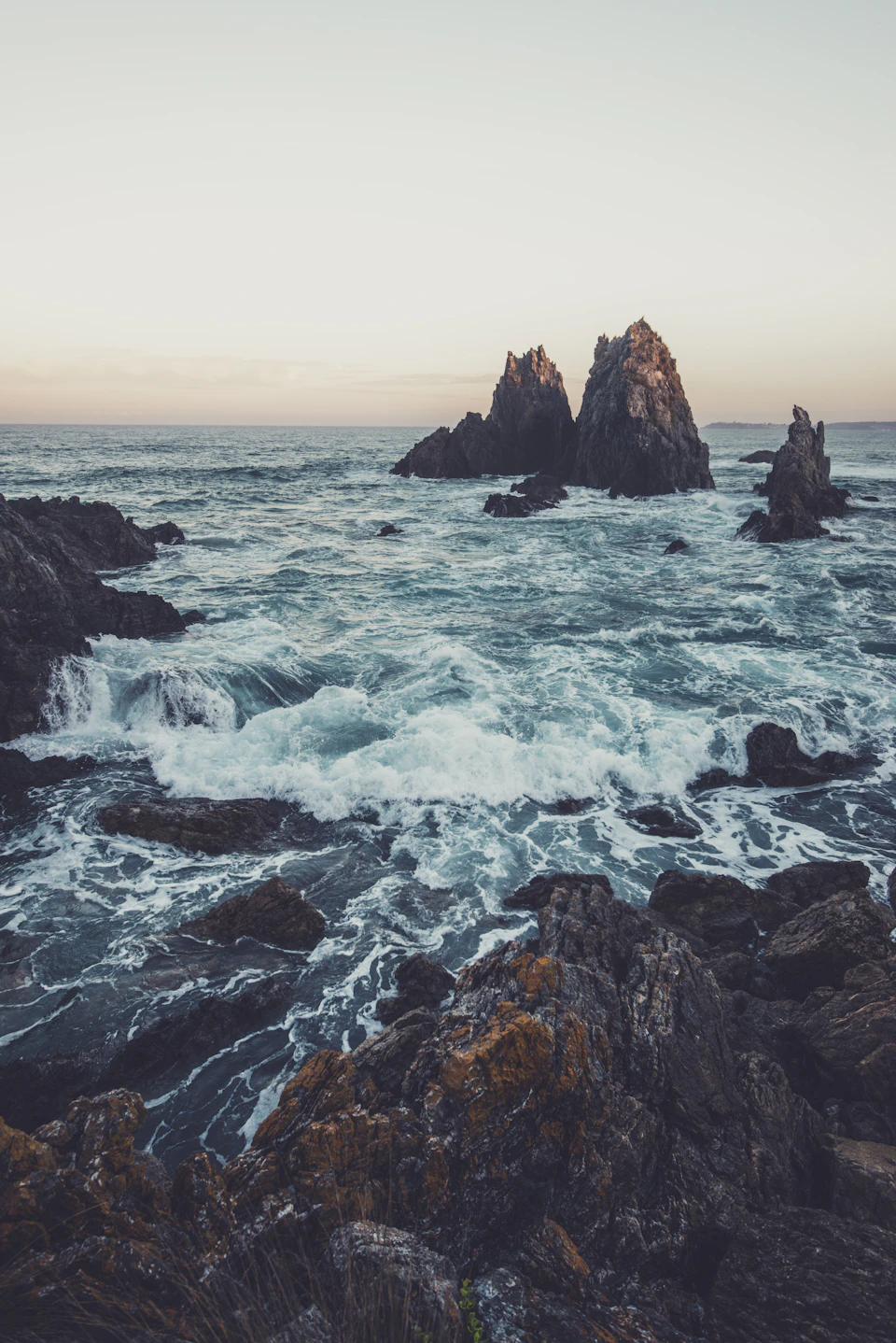 Two large, jagged rocks stand out on a rocky coastline with crashing ocean waves