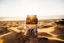 A can of Maui Brewing Co. beer is placed on a sandy beach with the ocean in the background. The sun is setting, casting a warm golden glow over the scene. The sand is textured and slightly uneven. The beer can features a wave design and tropical patterns.