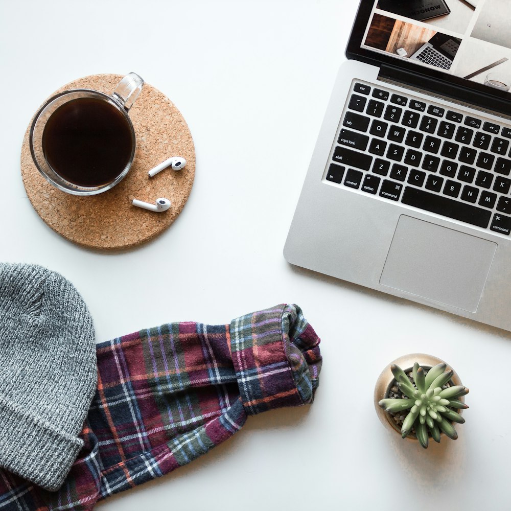 Foto ravvicinata di computer portatile acceso accanto a una tazza di caffè in vetro trasparente, una pianta grassa e un cappello lavorato a maglia grigio