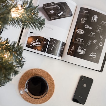 An open book displays pages with space-related illustrations and typography. The left page features the words 'The Space Exploration' alongside star constellations, while the right page showcases sketches of hands and technology, possibly related to space missions. Adjacent to the book, a cup filled with dark liquid rests on a cork coaster next to a smartphone, with a decorated tree partially visible in the top left corner, adding a festive ambience.