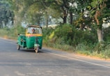 A green auto rickshaw is moving on a road lined with lush trees and dense vegetation. The driver is visible inside the vehicle, and the scene conveys a sense of motion with the blurred background.