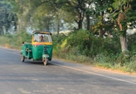 A green auto rickshaw is moving on a road lined with lush trees and dense vegetation. The driver is visible inside the vehicle, and the scene conveys a sense of motion with the blurred background.