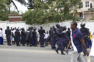 A group of uniformed police officers wearing protective gear are gathered near a crowd of people standing against a white wall. The officers appear to be communicating with the crowd. Trees and buildings are visible in the background, and a person in casual clothing walks by in the foreground.