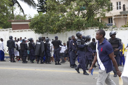 A group of uniformed police officers wearing protective gear are gathered near a crowd of people standing against a white wall. The officers appear to be communicating with the crowd. Trees and buildings are visible in the background, and a person in casual clothing walks by in the foreground.