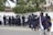 A group of uniformed police officers wearing protective gear are gathered near a crowd of people standing against a white wall. The officers appear to be communicating with the crowd. Trees and buildings are visible in the background, and a person in casual clothing walks by in the foreground.