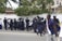 A group of uniformed police officers wearing protective gear are gathered near a crowd of people standing against a white wall. The officers appear to be communicating with the crowd. Trees and buildings are visible in the background, and a person in casual clothing walks by in the foreground.