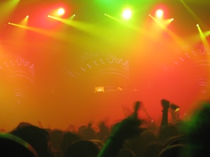 A vibrant and energetic concert or music festival scene, featuring a large crowd with raised hands silhouetted against a backdrop of colorful stage lights. The stage is partially visible with illuminated equipment and decorative patterns. The lights create a hazy atmosphere with predominant hues of red, yellow, and green.