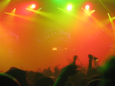 A vibrant and energetic concert or music festival scene, featuring a large crowd with raised hands silhouetted against a backdrop of colorful stage lights. The stage is partially visible with illuminated equipment and decorative patterns. The lights create a hazy atmosphere with predominant hues of red, yellow, and green.