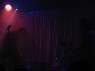 A dimly lit stage with silhouettes of musicians playing instruments. There's a visible microphone stand and a purple curtain in the background. A red spotlight shines from the top left, creating a moody atmosphere.