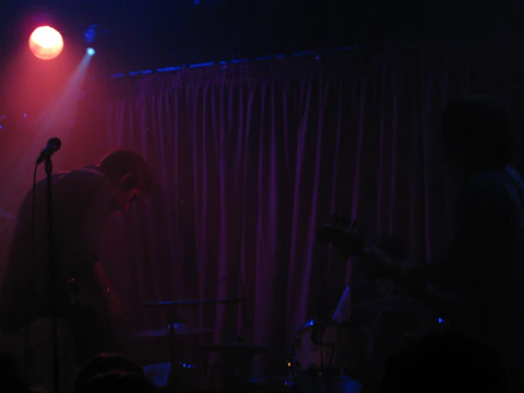 A dimly lit stage with silhouettes of musicians playing instruments. There's a visible microphone stand and a purple curtain in the background. A red spotlight shines from the top left, creating a moody atmosphere.