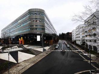 A modern office building with a curved facade, featuring large glass windows and multiple floors. Nearby, a street with parked cars extends into the distance, surrounded by other urban buildings. The sky appears overcast, and bare trees line the street.