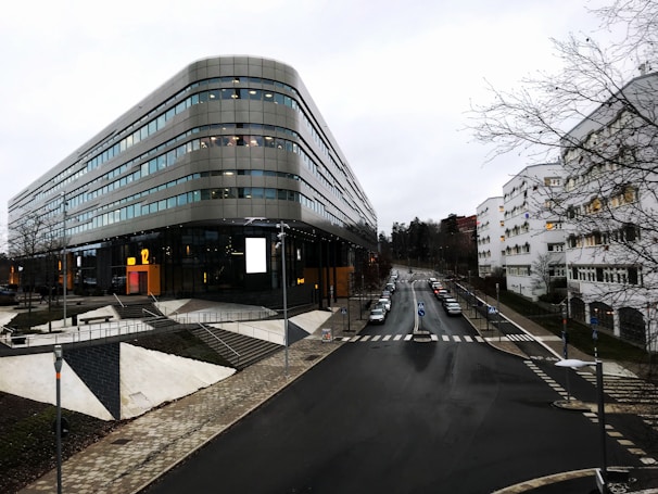 A modern office building with a curved facade, featuring large glass windows and multiple floors. Nearby, a street with parked cars extends into the distance, surrounded by other urban buildings. The sky appears overcast, and bare trees line the street.