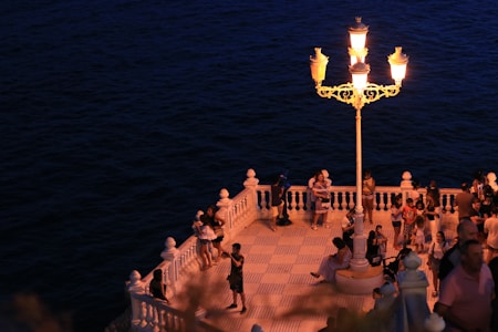 A group of people gather on a well-lit, tiled terrace by the sea. A decorative lamppost illuminates the scene, casting a warm glow. The terrace features white balustrades and overlooks a dark, expansive body of water. Some people are walking, others are interacting in small groups.