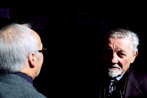 Two elderly men are partially illuminated by a strong light source against a dark background. One man is seen from the back, wearing a dark jacket and glasses. The other man's face is lit, showing his white hair and expressive, thoughtful expression as he looks towards the camera.