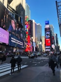 A bustling urban scene with numerous large digital billboards displaying various advertisements. Tall buildings surround the area, and people walk across the crosswalk. Vehicles are visible on the street, contributing to the lively city atmosphere under a clear blue sky.